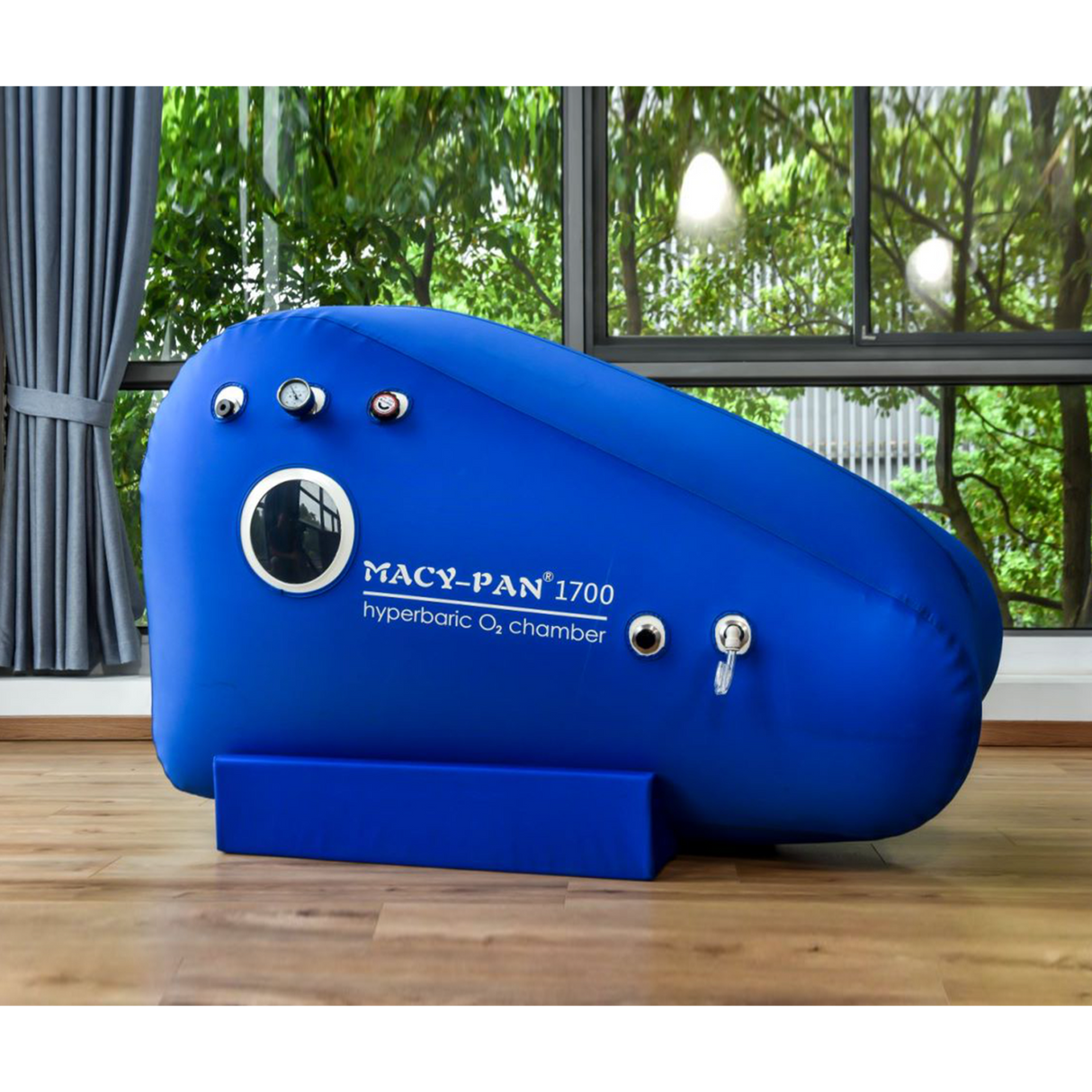 A blue Macy-Pan Hyperbaric Oxygen Therapy Chamber sitting type with visible pressure control valves and a logo on the side, placed in a room with a large window.